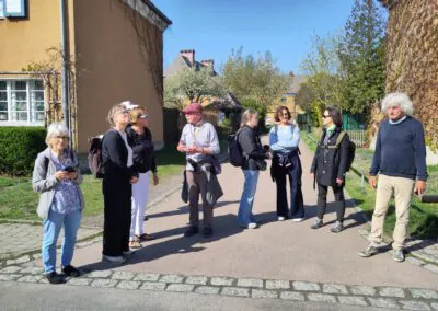 Eine Gruppe von neun Erwachsenen steht plaudernd auf einer sonnigen Straße, die von bunten Häusern und Bäumen gesäumt ist. Einige von ihnen halten Taschen in der Hand und tragen Sonnenbrillen oder Hüte, wobei die Schatten auf helles Tageslicht hinweisen.