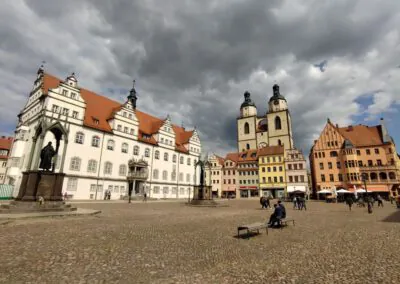 Ein großer kopfsteingepflasterter Platz in einer europäischen Stadt, umgeben von historischen Gebäuden mit roten Dächern und hohen Kirchtürmen; Menschen gehen spazieren und sitzen auf Bänken unter einem bewölkten Himmel.