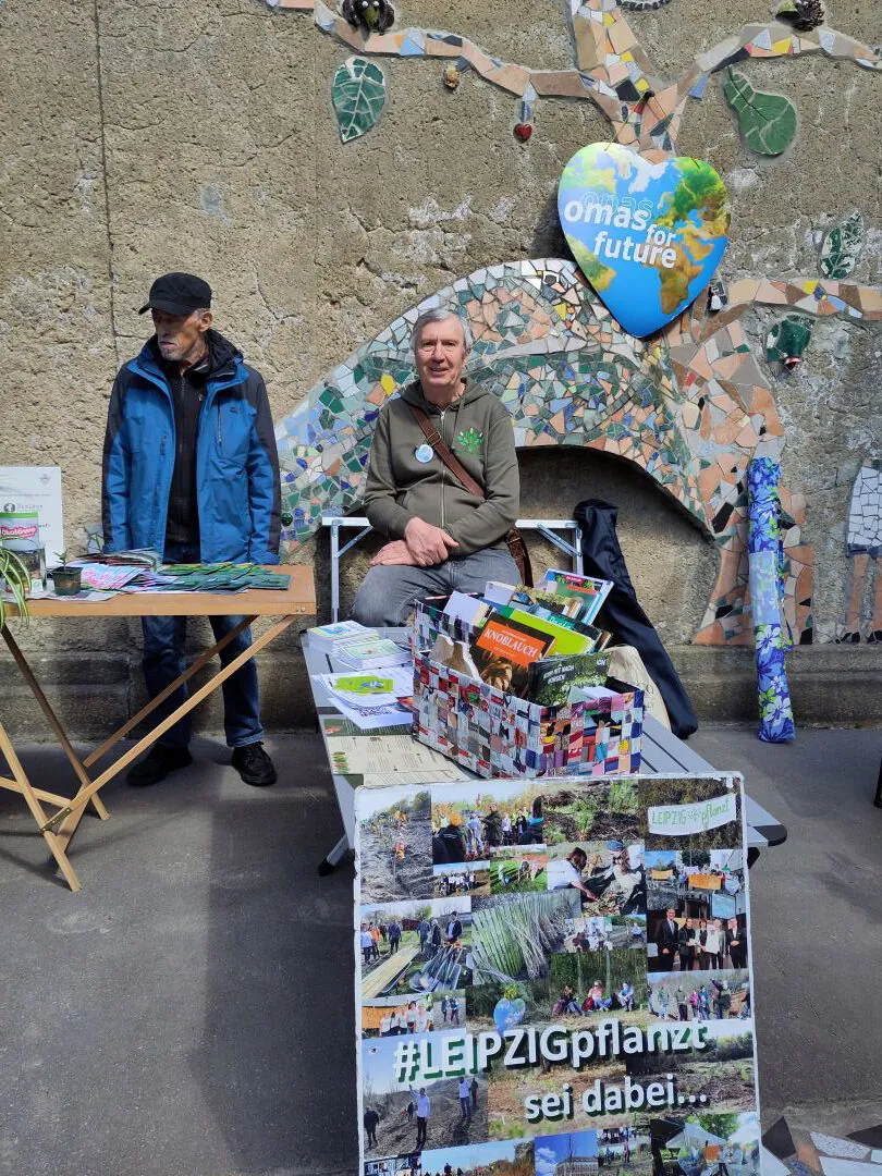 Zwei ältere Männer stehen und sitzen hinter einem Tisch mit Broschüren und Pflanzen bei einer Veranstaltung im Freien. Eine dekorierte Wand mit einem herzförmigen "omas for future"-Schild und eine Collagetafel mit der Aufschrift #LEIPZIGpflanze sind im Hintergrund zu sehen.