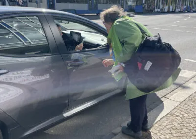 Eine Person in grüner Jacke und mit einer großen Tasche lehnt sich an das Fenster eines geparkten grauen Autos und spricht mit dem Fahrer in einer sonnigen Straße, in der im Hintergrund Straßenbahnen zu sehen sind.