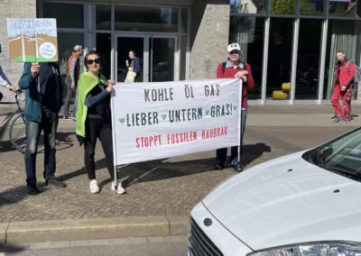 Drei Personen stehen auf einem Bürgersteig und halten ein Transparent mit der Aufschrift "Kohle Öl Gas lieber unterm Gras! Stoppt fossilen Raubbau" bei einer Protestaktion. Ein weiteres Schild in deutscher Sprache wird in der Nähe gehalten. Im Vordergrund ist ein weißes Auto zu sehen.