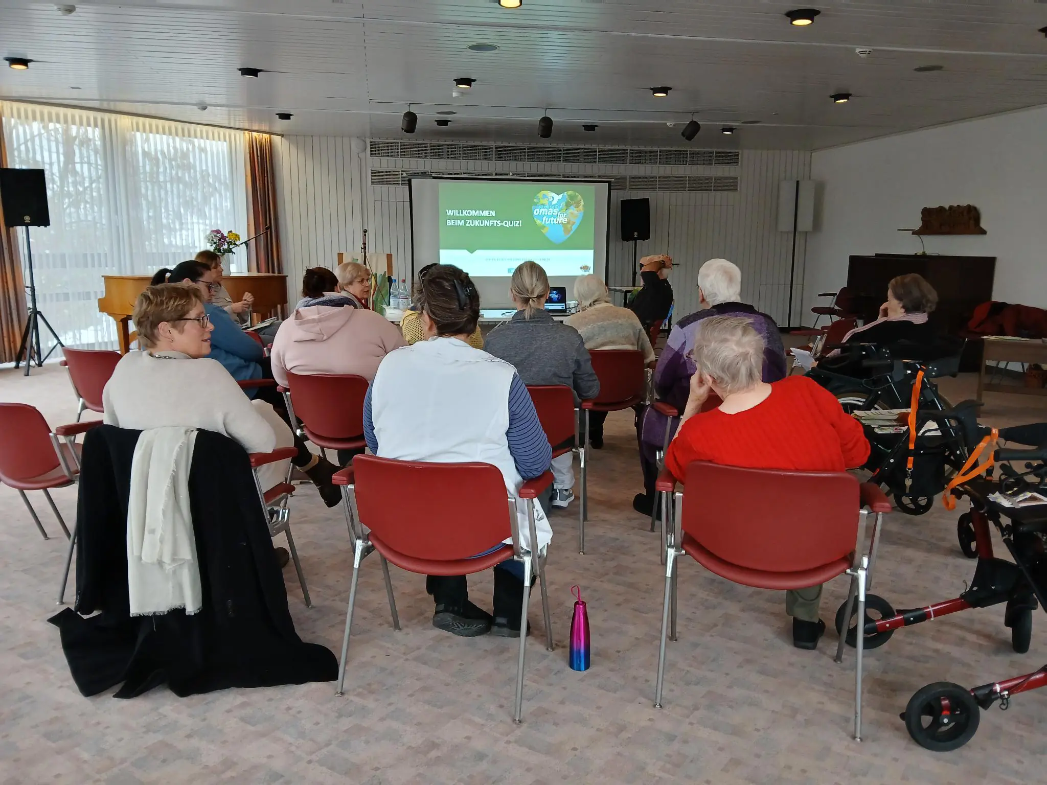 Eine Gruppe von Personen sitzt auf roten Stühlen in einem hellen Raum vor einer Projektionsfläche. Auf der Leinwand wird eine Begrüßungsbotschaft für ein Seniorencafé angezeigt. Einige Teilnehmer benutzen Gehhilfen; Mäntel hängen an den Stühlen in der Nähe.
