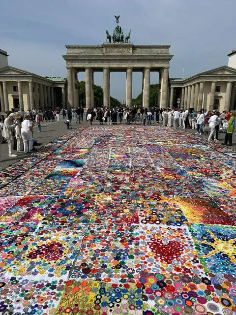 Ein farbenfroher Patchwork-Quilt bedeckt den Boden vor dem Brandenburger Tor in Berlin. Die Menschen haben sich versammelt und bewundern das Kunstwerk unter freiem Himmel.