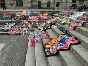 Bunte gehäkelte Decken und Stücke mit Blumenmustern sind auf breiten Steinstufen im Freien ausgestellt und bilden einen lebhaften Kontrast zu den grauen Stufen und dem Bürgersteig. Im Hintergrund sind Fahrräder und ein Gebäude zu sehen.