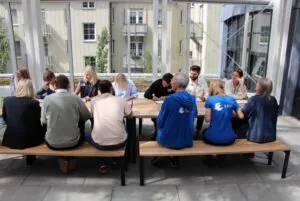 Eine Gruppe von Menschen sitzt an einem langen Holztisch im Freien, einige mit dem Gesicht zueinander, andere mit dem Rücken zur Kamera. Zwei Personen tragen blaue Hemden mit weißen Logos. Im Hintergrund sind Gebäude und Grünflächen zu sehen.
