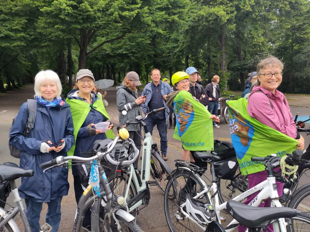 Eine Gruppe von Menschen, überwiegend ältere Erwachsene, steht mit Fahrrädern in einem Park. Vier Frauen im Vordergrund lächeln und tragen grüne Umhänge oder Westen. Im Hintergrund sind unter bewölktem Himmel Bäume und weitere Teilnehmer zu sehen.