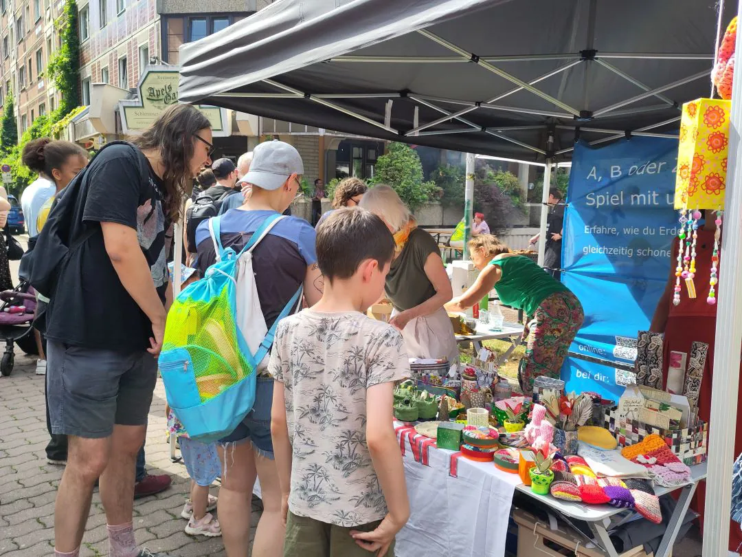 Eine Gruppe von Menschen, darunter Kinder und Erwachsene, begutachtet handgefertigte Kunsthandwerksartikel und bunte Waren, die auf Tischen unter dem Vordach eines Marktstandes auf einem Straßenfest im Freien ausgestellt sind. Im Hintergrund ist ein blaues Informationsbanner zu sehen.