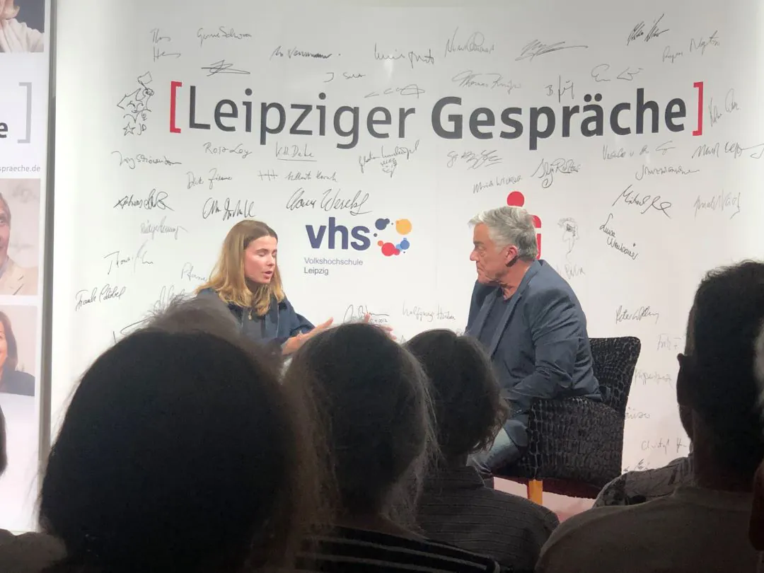 Auf einer Bühne im Rahmen der Veranstaltung „Leipziger Gespräche“ sitzen zwei Personen und unterhalten sich. Im Hintergrund sind zahlreiche Unterschriften und das Logo der vhs Leipzig zu sehen. Im Vordergrund sind Zuschauer zu sehen.