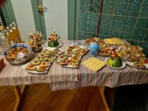 4.10.26 5 Jahre O4F Bretten©O4 - Omas for Future Auf einem langen Tisch, der mit einer gestreiften Tischdecke bedeckt ist, stehen verschiedene Häppchen, darunter kleine Sandwiches, Obstspieße und eine Schüssel mit frittierten Snacks.