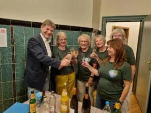 5 Jahre O4F 4.10.26 Bretten©O4 - Omas for Future Ein Mann im Anzug und fünf Frauen in einheitlichen grünen Shirts stoßen mit Gläsern Sekt an.