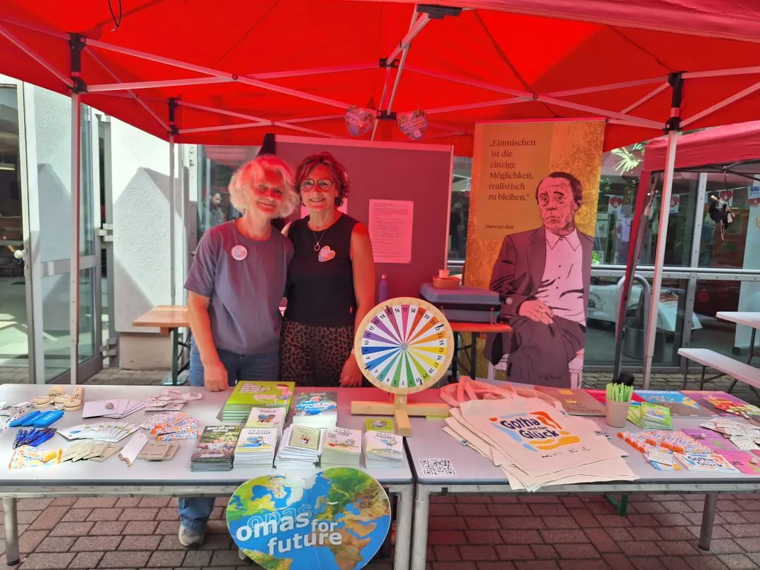 Hinter einem Stand mit Büchern, Flyern und einem Spinnrad unter roten Zelten stehen zwei Frauen. Hinter ihnen steht eine Pappfigur eines Mannes. Auf dem Tisch liegen Materialien von „Omas for Future“.